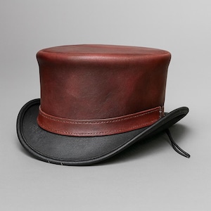May include: A burgundy leather top hat with a black brim and a brown band. The hat has a classic, cylindrical crown and a curved brim. The hat is a formal accessory.