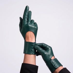 May include: A pair of dark forest green leather gloves featuring a cutout design on the back of the hand and a wide wrist cuff secured with two matching green snap buttons. The smooth leather material covers the fingers and palm area.