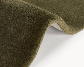 May include: Close-up of a section of a green rug. The rug has a soft, plush texture and a deep olive colour. The edge of the rug is visible, revealing a lighter backing. The rug is folded over, showing the texture and colour variations.