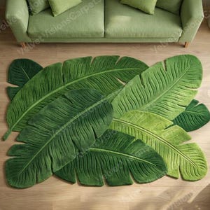 May include: A green rug in the shape of large banana leaves. The rug has a soft, textured surface with detailed veining. It's placed on a wooden floor, with a green sofa and plants visible in the background.