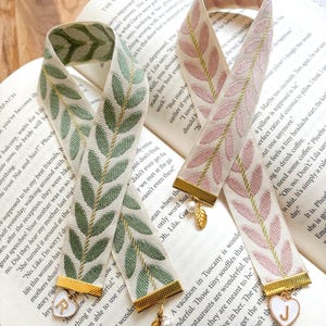 Op de afbeelding: Twee decoratieve bladwijzers met bladpatronen in groen en roze, gouden randen en bedels. E&eacute;n bladwijzer heeft een gouden hartbedel met de letter "R", de andere met de letter "J".