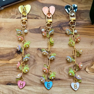 May include: Three decorative keychains with heart-shaped clasps and charms. Each keychain features a gold chain adorned with colorful butterfly and flower charms. The bottom charms are heart-shaped with the letters H, E, and B.