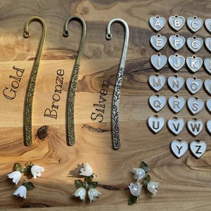 May include: A selection of decorative bookmarks in gold, bronze, and silver, each with a floral pattern. Also shown are heart-shaped charms with letters A-Z, and small white flower accents.