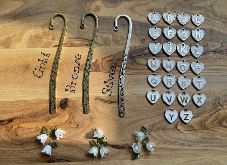 May include: A selection of decorative bookmarks in gold, bronze, and silver, each with a floral pattern. Also shown are heart-shaped charms with letters A-Z, and small white flower accents.