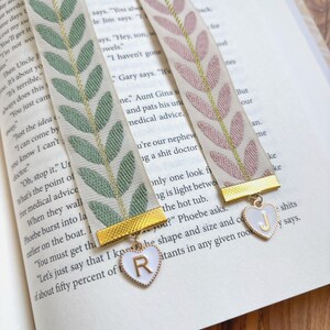 Op de afbeelding: Twee decoratieve lint boekenleggers met bladpatronen in groen en roze, geaccentueerd met gouden afwerking. Elke boekenlegger heeft een hartvormige bedel met een letter, rustend op de pagina's van een boek.