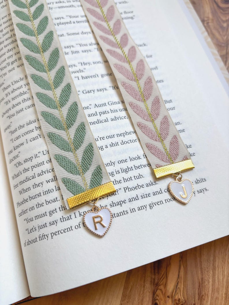 Op de afbeelding: Twee decoratieve lint boekenleggers met bladpatronen in groen en roze, geaccentueerd met gouden afwerking. Elke boekenlegger heeft een hartvormige bedel met een letter, rustend op de pagina's van een boek.