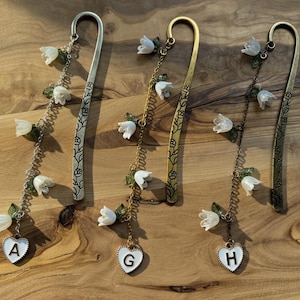 May include: Three metal bookmarks with floral and heart charms. The bookmarks are silver, gold, and bronze. Each bookmark has a chain with white flower and green leaf accents. The charms are white hearts with the letters A, G, and H.