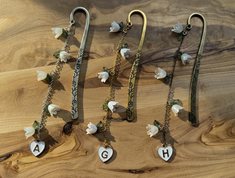May include: Three metal bookmarks with floral and heart charms. The bookmarks are silver, gold, and bronze. Each bookmark has a chain with white flower and green leaf accents. The charms are white hearts with the letters A, G, and H.