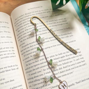 May include: A decorative gold bookmark with a floral pattern and a chain of white flower charms. The chain ends with a white heart pendant with the letter "H". The bookmark is on the pages of a book.