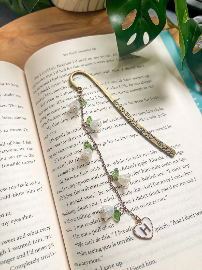 May include: A decorative gold bookmark with a floral pattern and a chain of white flower charms. The chain ends with a white heart pendant with the letter "H". The bookmark is on the pages of a book.
