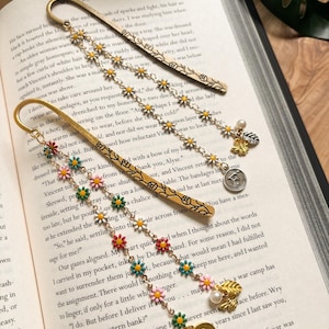 May include: Three decorative metal bookmarks with daisy chains and charms rest on an open book. The bookmarks feature colorful daisy accents, a bee charm, a pearl, and a coin-shaped charm with a letter.