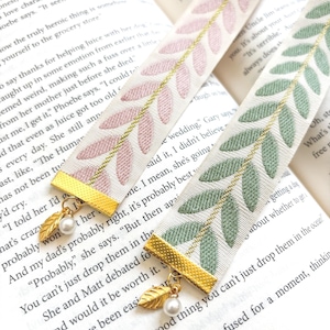 Op de afbeelding: Twee decoratieve lint bladwijzers met bladeren patronen. E&eacute;n bladwijzer heeft roze bladeren, de andere groene. Beide hebben goudkleurige metalen accenten met een blad charme en een parel.