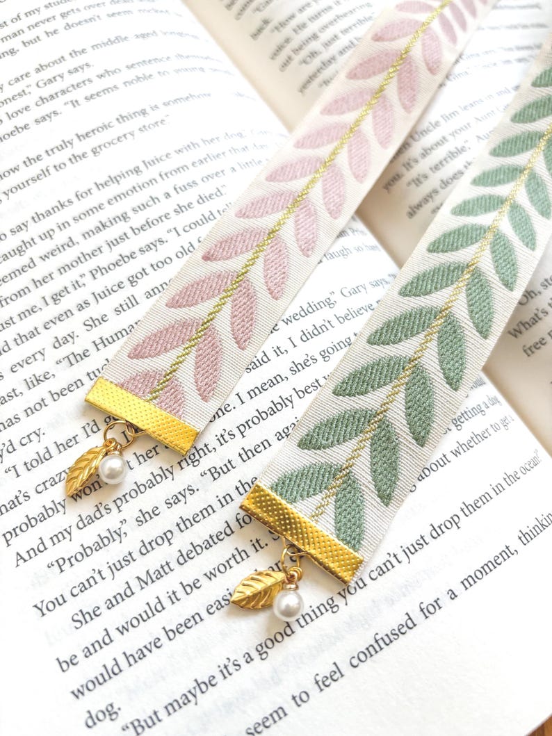 Op de afbeelding: Twee decoratieve lint bladwijzers met bladeren patronen. E&eacute;n bladwijzer heeft roze bladeren, de andere groene. Beide hebben goudkleurige metalen accenten met een blad charme en een parel.