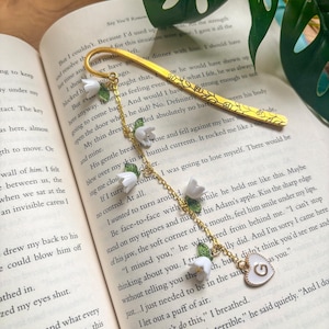 May include: A gold bookmark with a floral design and a heart-shaped charm. The bookmark features white flower and green leaf accents, and is resting on the pages of a book.