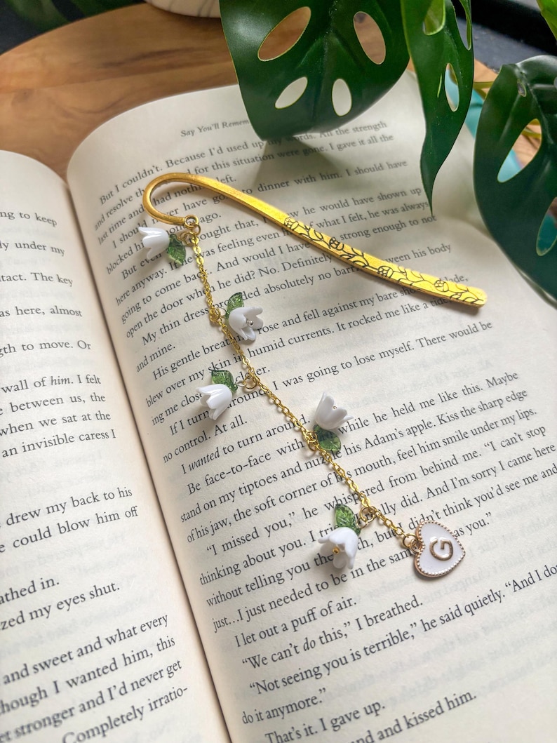 May include: A gold bookmark with a floral design and a heart-shaped charm. The bookmark features white flower and green leaf accents, and is resting on the pages of a book.