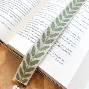 Op de afbeelding: Decoratieve boekenlegger met een cr&egrave;mekleurige achtergrond en een groen bladerpatroon. De boekenlegger heeft een goudkleurig metalen uiteinde met een wit hartvormig bedeltje met de letter "R". De boekenlegger ligt in een open boek.