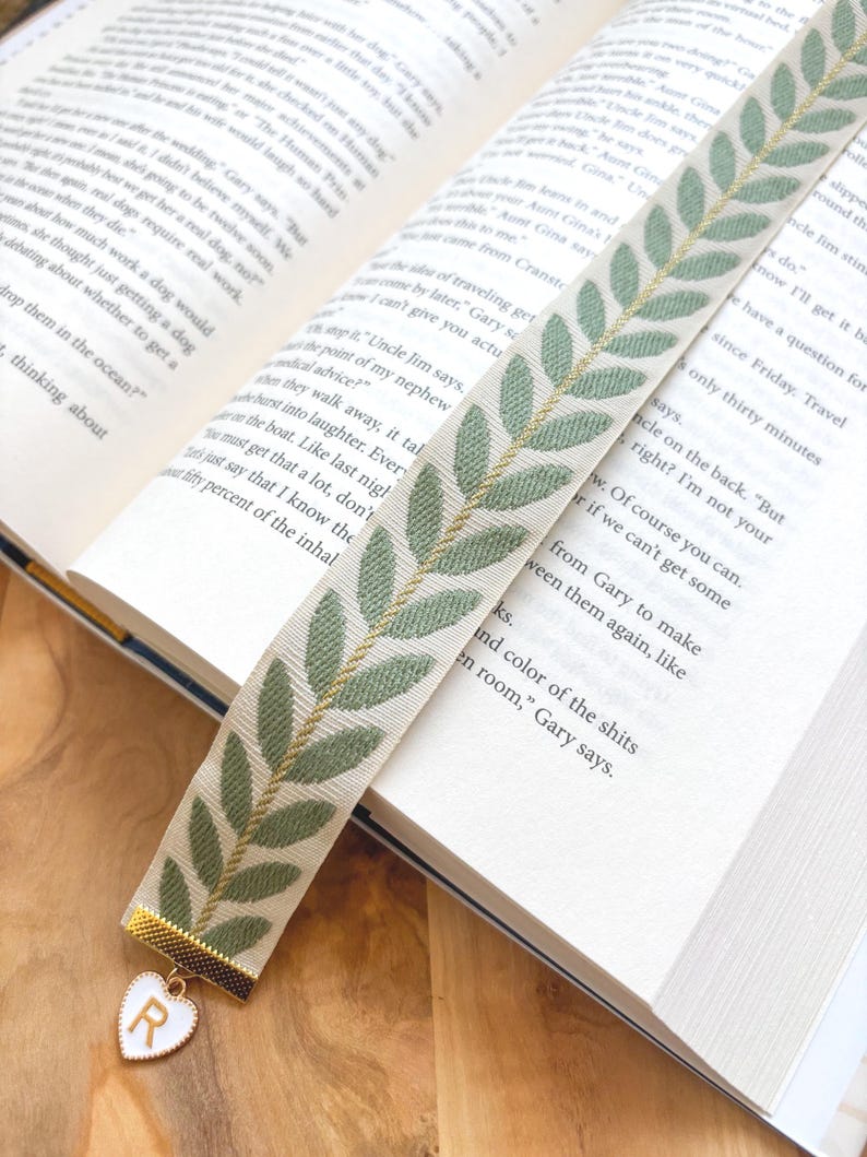 Op de afbeelding: Decoratieve boekenlegger met een cr&egrave;mekleurige achtergrond en een groen bladerpatroon. De boekenlegger heeft een goudkleurig metalen uiteinde met een wit hartvormig bedeltje met de letter "R". De boekenlegger ligt in een open boek.