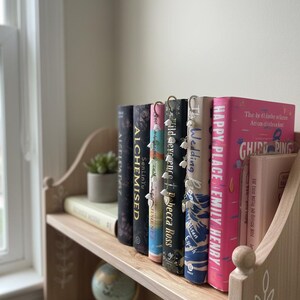 May include: A light wooden bookshelf displaying a collection of books. The books have different coloured spines and visible titles. Some books have decorative bookmarks. A small potted plant and a miniature globe are also on the shelf.