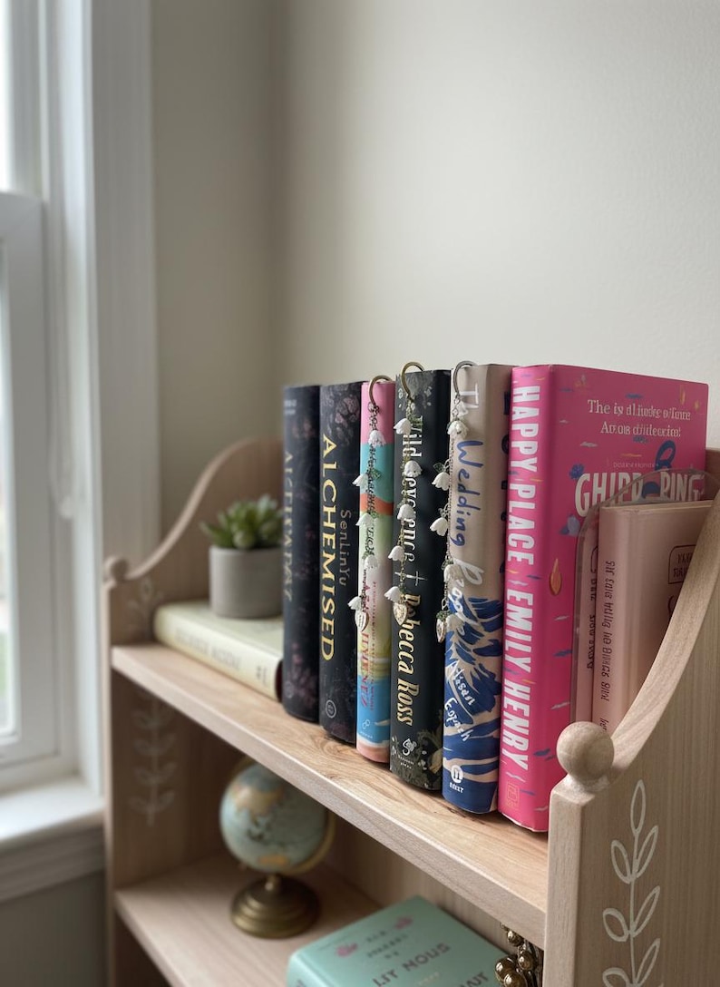 May include: A light wooden bookshelf displaying a collection of books. The books have different coloured spines and visible titles. Some books have decorative bookmarks. A small potted plant and a miniature globe are also on the shelf.