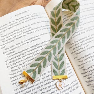 Op de afbeelding: Een decoratieve bladwijzer met een groen bladerpatroon op een cr&egrave;mekleurige achtergrond. De bladwijzer heeft gouden accenten en een bedel met de letter "R" op een hart. De bladwijzer ligt op de pagina's van een boek.