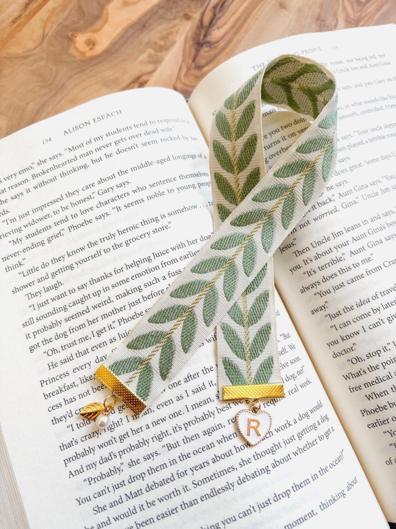 Op de afbeelding: Een decoratieve bladwijzer met een groen bladerpatroon op een cr&egrave;mekleurige achtergrond. De bladwijzer heeft gouden accenten en een bedel met de letter "R" op een hart. De bladwijzer ligt op de pagina's van een boek.