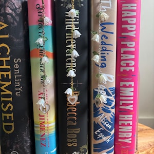 May include: Books with floral bookmarks. Each bookmark has a chain with white flower charms and a heart charm. The book spines are various colours, with titles and author names visible.