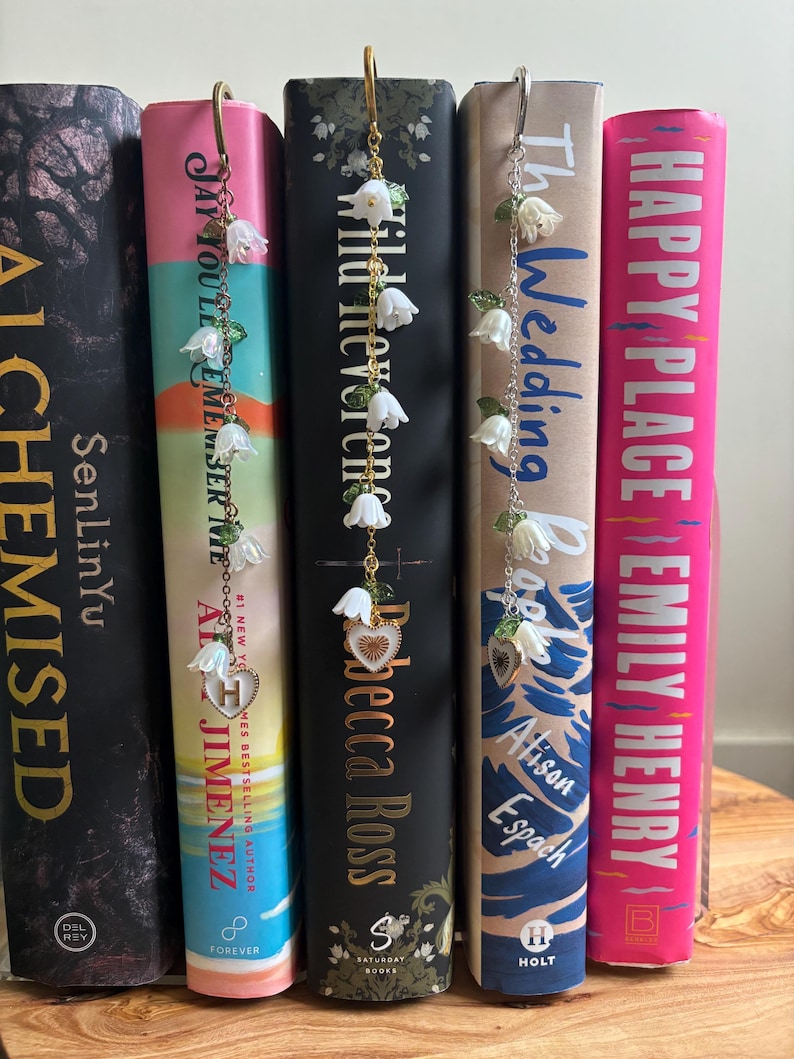 May include: Books with floral bookmarks. Each bookmark has a chain with white flower charms and a heart charm. The book spines are various colours, with titles and author names visible.