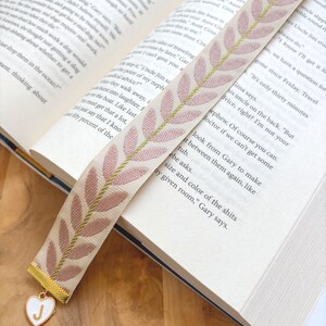 Op de afbeelding: Een stoffen bladwijzer met een cr&egrave;mekleurige basis en een patroon van roze bladeren en gouden accenten. Een goudkleurige bedel met een wit hart en de letter "J" hangt aan de onderkant. De bladwijzer ligt in een open boek.