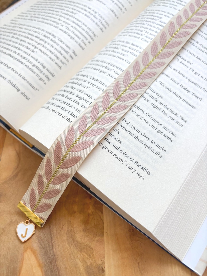 Op de afbeelding: Een stoffen bladwijzer met een cr&egrave;mekleurige basis en een patroon van roze bladeren en gouden accenten. Een goudkleurige bedel met een wit hart en de letter "J" hangt aan de onderkant. De bladwijzer ligt in een open boek.