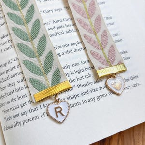 Op de afbeelding: Twee decoratieve bladwijzers met bladpatronen in groen en roze, met gouden accenten. Elke bladwijzer heeft een goudkleurige metalen sluiting en een hartvormige bedel met een letter. De bladwijzers liggen op de pagina's van een boek.