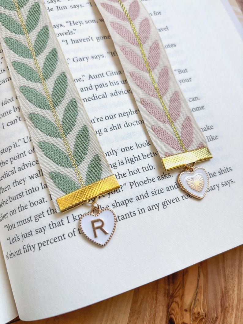 Op de afbeelding: Twee decoratieve bladwijzers met bladpatronen in groen en roze, met gouden accenten. Elke bladwijzer heeft een goudkleurige metalen sluiting en een hartvormige bedel met een letter. De bladwijzers liggen op de pagina's van een boek.