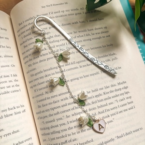 May include: A silver bookmark with a floral pattern and a hanging chain. The chain features white flower charms with green leaves and a heart-shaped charm with the letter "A". The bookmark is placed on the open pages of a book.