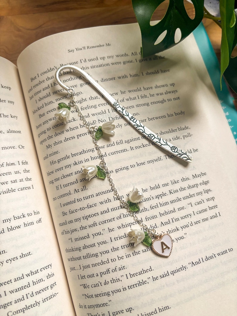 May include: A silver bookmark with a floral pattern and a hanging chain. The chain features white flower charms with green leaves and a heart-shaped charm with the letter "A". The bookmark is placed on the open pages of a book.