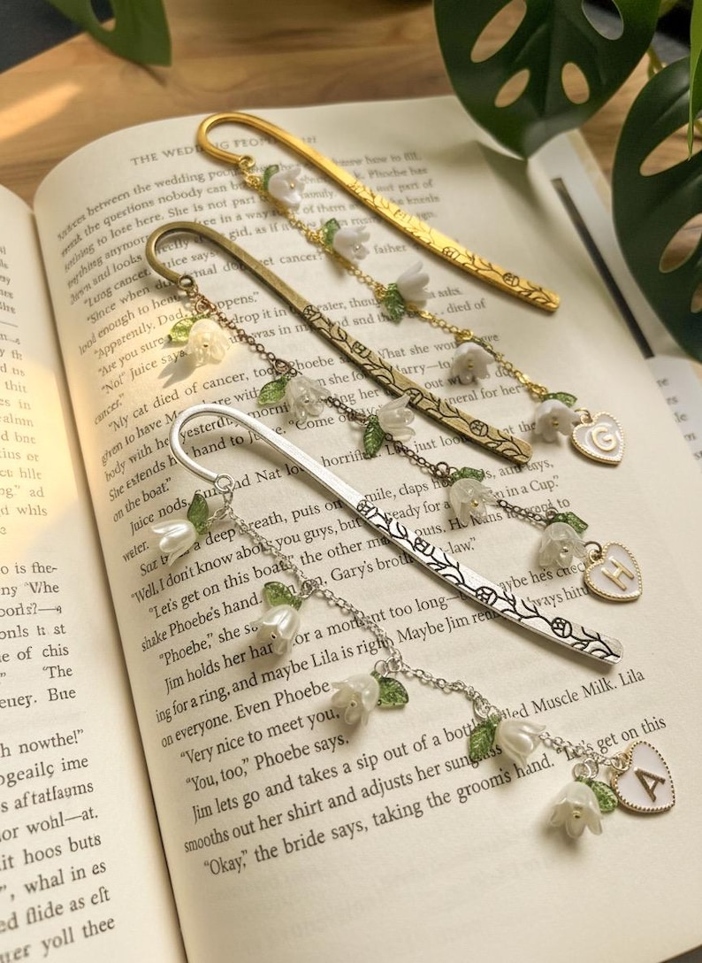 May include: Four metal bookmarks with floral and initial charms on an open book. The bookmarks are gold, bronze, and silver, each with white flowers, green leaves, and a charm with a letter.