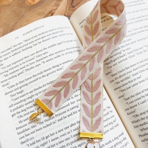 Op de afbeelding: Cr&egrave;mekleurige bladwijzer met roze bladeren en gouden accenten. Een witte hartbedel met de letter "J" is bevestigd. De bladwijzer ligt in een open boek.