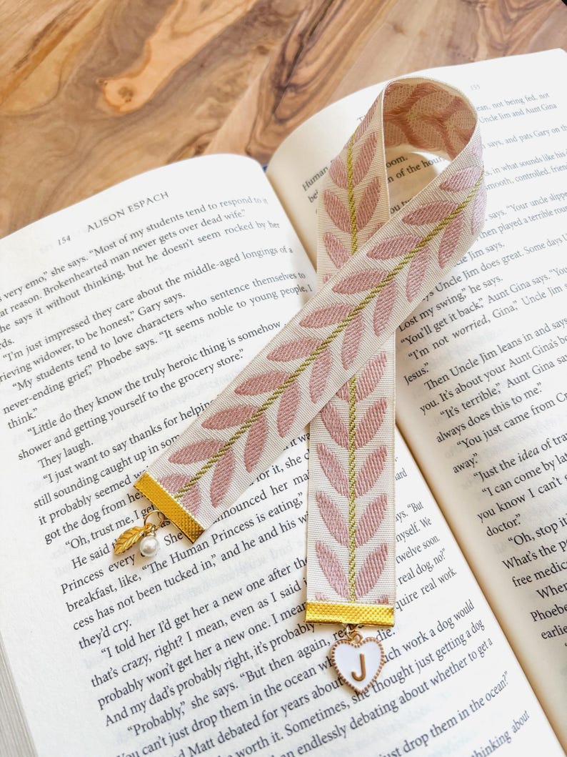 Op de afbeelding: Cr&egrave;mekleurige bladwijzer met roze bladeren en gouden accenten. Een witte hartbedel met de letter "J" is bevestigd. De bladwijzer ligt in een open boek.