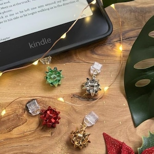 May include: A wooden surface displays four bow-shaped earrings in red, green, silver, and gold. A Kindle e-reader, string lights, and Christmas decorations are also present. The word "kindle" is visible on the e-reader.