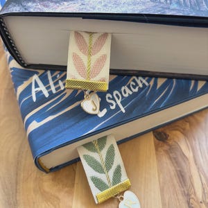 Op de afbeelding: Twee decoratieve bladwijzers met bloemmotieven zijn in boeken geplaatst. E&eacute;n bladwijzer heeft een roze bloemmotief, de andere een groen bloemmotief. Beide hebben gouden randen en hartvormige bedels met initialen.
