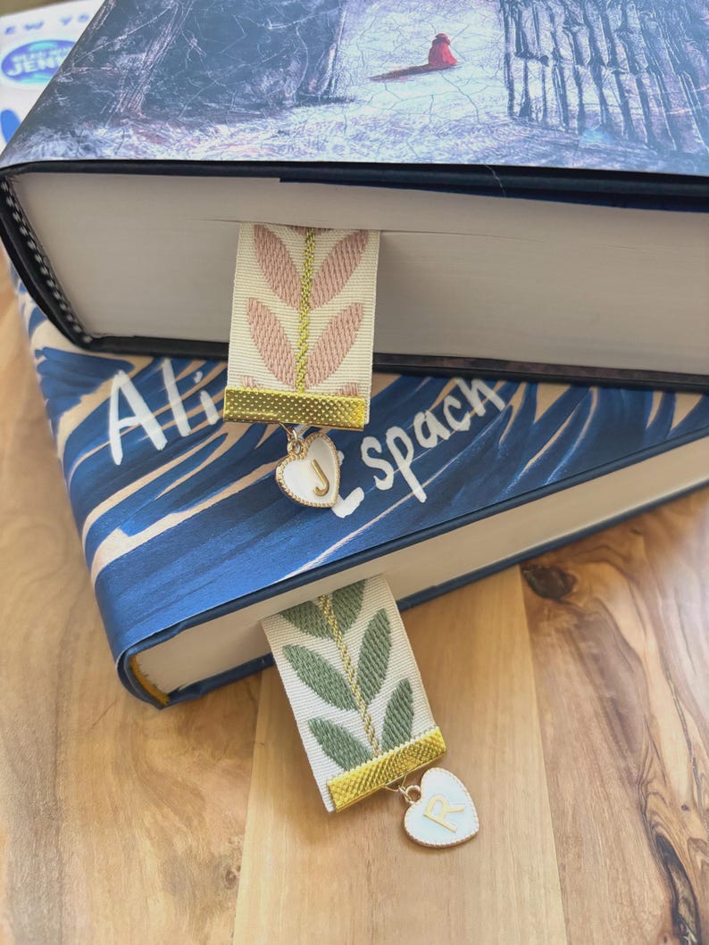 Op de afbeelding: Twee decoratieve bladwijzers met bloemmotieven zijn in boeken geplaatst. E&eacute;n bladwijzer heeft een roze bloemmotief, de andere een groen bloemmotief. Beide hebben gouden randen en hartvormige bedels met initialen.
