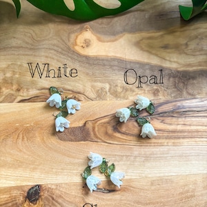 May include: Three pairs of floral earrings in white, opal, and clear. Each pair features small white flower clusters with green leaves. The earrings are displayed on a wooden surface, with the words "White", "Opal", and "Clear" written below each pair.