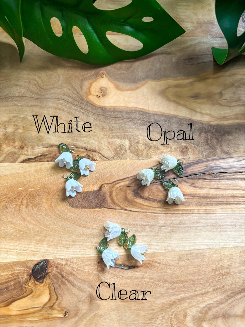 May include: Three pairs of floral earrings in white, opal, and clear. Each pair features small white flower clusters with green leaves. The earrings are displayed on a wooden surface, with the words "White", "Opal", and "Clear" written below each pair.