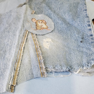 May include: A light blue denim patch with a frayed edge and a small, embroidered mouse applique. The mouse is tan and brown, with a smiling face. The denim has a distressed look.