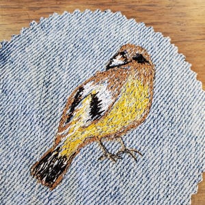 Puede incluir: Pájaro bordado sobre un parche de mezclilla. El pájaro está bordado con hilo amarillo, marrón, negro y blanco. El parche de mezclilla tiene un borde festoneado. Fondo de madera.