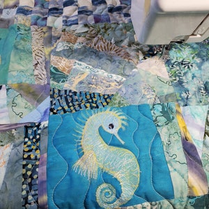 以下が含まれることがあります： 海をテーマにしたキルトで、タツノオトシゴと魚の刺繍が施されています。キルトは、さまざまな青と緑の布のパッチで構成されており、ミシン針が右上に見えます。