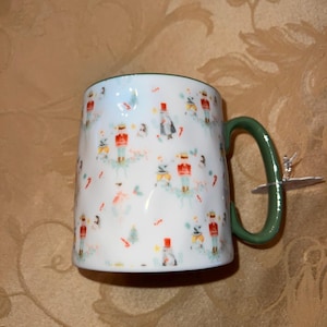 May include: White ceramic mug with a green handle and rim. The mug is decorated with a festive pattern of nutcrackers, snowmen, and Christmas trees. A perfect gift for the holiday season.