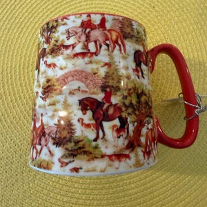 May include: A white ceramic mug with a red handle featuring a detailed wrap-around illustration of a fox hunt. The scene depicts riders on horseback, hunting dogs, trees, and a stone bridge in shades of brown, green, and red against a white background.