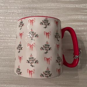 May include: White ceramic mug with a red interior and handle. The mug is decorated with a pattern of Christmas trees and pink bows. The handle has two rings on it.