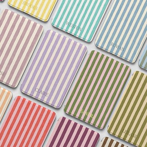 Puede incluir: Una colección de estuches rectangulares con rayas verticales en varios colores, incluyendo amarillo, azul verdoso, azul, rosa, morado, verde y naranja. Cada estuche está personalizado con un nombre impreso en una fuente sencilla, como Brooks, Weston, Rowan, Claire, Eloise, Sandr y Leilani.
