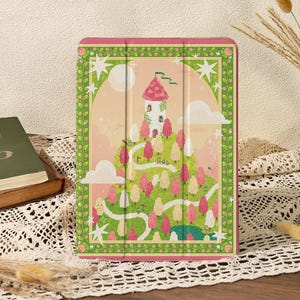 Puede incluir: Una funda rectangular para tableta con una ilustración caprichosa de una torre con techo rosa sobre una colina verde. La escena incluye árboles rosas y blancos, un camino sinuoso, nubes blancas y estrellas, enmarcado por un borde floral verde decorativo.