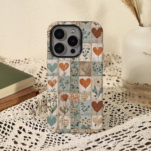 Puede incluir: Funda de teléfono estampada con un diseño de cuadrícula. Presenta corazones y motivos florales en tonos naranja, azul y marrón. La funda tiene un recorte para la cámara y un acabado brillante.